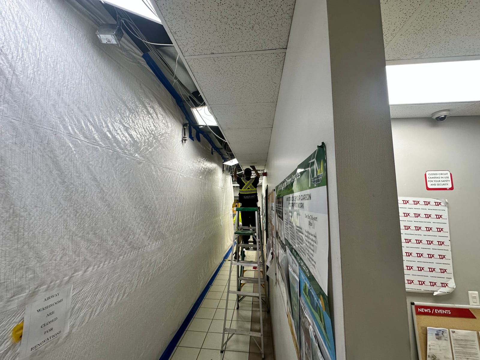 Worker installing T-bar ceiling tiles from a ladder in a commercial hallway